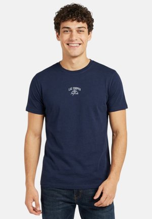 Jeune homme souriant portant un t-shirt Lee Cooper bleu marine avec un petit logo blanc et un jean foncé, debout devant un fond clair uni.