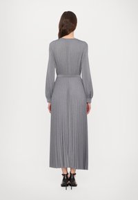 Robe plissée grise à manches longues et taille cintrée, avec une texture lisse. Portée avec des chaussures à talons hauts noires.