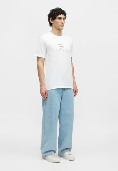Νέος άνδρας με σγουρά μαλλιά που φοράει ένα λευκό T-shirt με την επιγραφή «Limited Edition», ανοιχτό μπλε φαρδιά τζιν και λευκά αθλητικά παπούτσια, που στέκεται μπροστά σε λευκό φόντο.