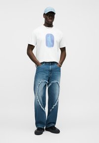 Λευκό γραφικό t-shirt με μπλε σχέδιο· τζιν παντελόνι με φθορές και σχισίματα σε σχήμα καρδιάς· ανοιχτό μπλε καπέλο· μαύρα παπούτσια· ασημένια αλυσίδα.