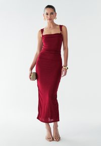Červené priliehavé šaty so štvorcovým výstrihom, nariasenou textúrou a širokými ramienkami. Dopĺňa ich obuv na remienkoch a malá kabelka.