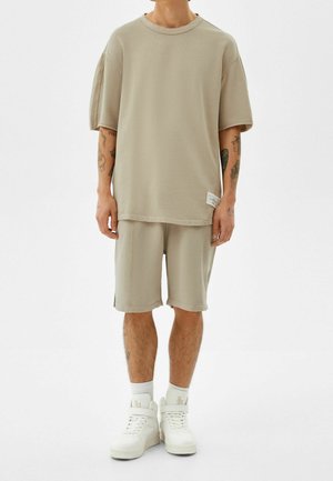 Maglietta oversized in cotone beige con maniche corte, abbinata a pantaloni corti coordinati. Entrambi presentano una vestibilità comoda e dettagli di cucitura minimi.