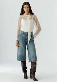 Vrouw in een doorschijnende witte kanten blouse met strik, denim korte broek tot de knie en donkere leren laarzen tot de knie, staand tegen een effen achtergrond.