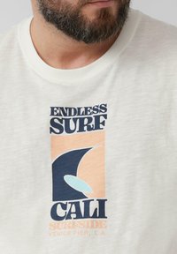 Witte katoenen t-shirt met een grafisch ontwerp van "ENDLESS SURF," een donkere blauwe golf en pastelaccenten. Ronde hals, ontspannen pasvorm.