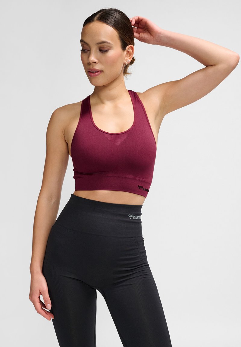 Femme portant un soutien-gorge de sport bordeaux et un legging noir taille haute, posant avec un bras levé et regardant vers le bas.