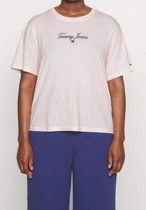 Henkilö, joka käyttää vaaleanpunaista "Tommy Jeans" -T-paitaa ja tummansinisiä housuja, näkyy ylävartalo ja kädet yksiväristä taustaa vasten.