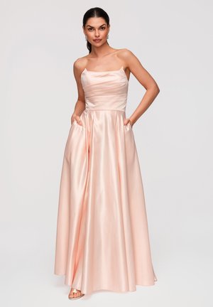 Vrouw staat met handen in de zakken en draagt een strapless, satijnen, blush roze avondjurk met een aansluitend lijfje en een volle rok.