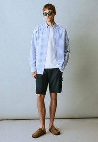 Giovane uomo con occhiali da sole, una camicia a righe sopra una maglietta bianca, pantaloni corti neri e scarpe slip-on marroni, si trova in un ambiente sfocato.