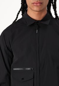Black lightweight jacket with a full zip, collar, and a chest pocket featuring a zip closure and a flap pocket with a snap button.