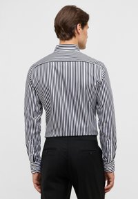Camicetta a righe in bianco e nero, con maniche lunghe, colletto classico e polsini con bottoni, dal taglio sartoriale. I pantaloni sono neri.