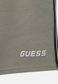 Harmaa kangas lyhyet housut, joissa on sileä rakenne, mustat sivuviivat ja selkeä hopeinen "GUESS"-logo helman kohdalla.