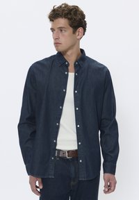 Camisa de manga larga de denim oscuro con cuello abotonado, botones metálicos y un ajuste relajado, combinada con una camiseta sin mangas blanca de canalé y jeans azules.