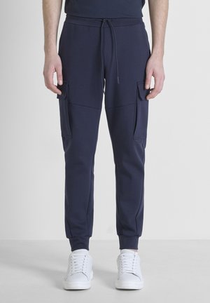 Pantalones jogger azul marino hechos de una tela suave, con cintura elástica, cordón, bolsillos de carga laterales y puños en los tobillos ajustados. Combinados con zapatillas blancas.