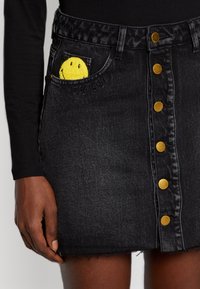 Gonna in denim nera con chiusura a bottoni frontale, orlo sfilacciato e un dettaglio a tasca con faccina sorridente gialla e il testo "LA GIOIA È ADESSO."