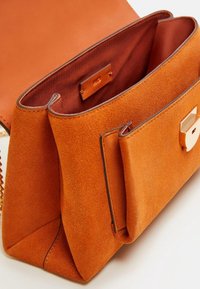 Sac à main en daim orange avec une finition texturée, doté d'une bandoulière chaîne et d'une fermeture à rabat dorée. L'intérieur est doublé d'un tissu plus sombre.