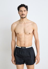 Zwarte boxershorts met een knoopsluiting, voorzien van een contrasterende tailleband met het label "BOSS" in zwart en grijs. Gladde textuur en casual pasvorm.
