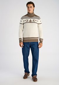 Stickad polotröja i cremefärg med bruna geometriska mönster och renmotiv, kombinerad med blå jeans och bruna skor.