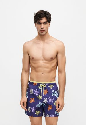 Homme aux cheveux foncés portant un short de bain violet avec des imprimés colorés de tortues, debout devant un fond clair uni.