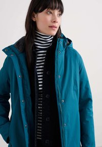 Giacca impermeabile turchese con cappuccio, bottoni a pressione argentati e un morbido interno grigio. Indossata sopra una camicia di jeans nera e un dolcevita a righe.