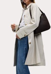 Lichtbeige trenchcoat met grote ronde knopen, gecombineerd met een witte top en blauwe denim jeans. Een donkerbruine, gestructureerde handtas wordt gedragen.