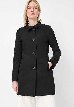 Woman wearing a fitted black buttoned coat with collar and long sleeves, standing with one hand in pocket against white background.