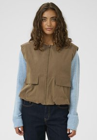 Kvinde med skulderlangt krøllet hår iført en brun ærmeløs zipvest over en lys blå strikket langærmet sweater og mørke jeans.