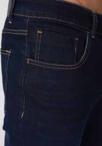 Σκούρο τζιν με πορτοκαλί ραφές αντίθεσης, δομημένες τσέπες και λεία υφή. Η εφαρμογή είναι εφαρμοστή γύρω από τη μέση και τους μηρούς.