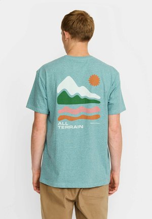 Joven visto desde atrás con camiseta azul verdosa con gráfico de montaña, sol y olas y texto "ALL TERRAIN", combinado con pantalones color beige.