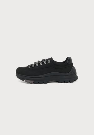 Μαύρο χαμηλό sneaker με παχιά λαστιχένια σόλα, μεταλλικά κορδόνια και στρογγυλά μαύρα-άσπρα κορδόνια, πλευρική όψη σε λευκό φόντο.