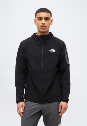 Νεαρός άνδρας που φοράει μια μαύρη ζακέτα North Face με φερμουάρ και κουκούλα, καθώς και γκρι παντελόνι, στέκεται μπροστά σε απλό φωτεινό φόντο.