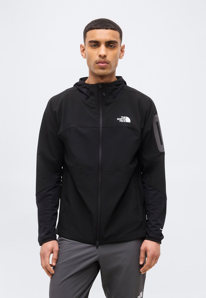 Νεαρός άνδρας που φοράει μια μαύρη ζακέτα North Face με φερμουάρ και κουκούλα, καθώς και γκρι παντελόνι, στέκεται μπροστά σε απλό φωτεινό φόντο.