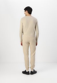 Beige langermet topp og matchende joggebukser, begge i et mykt, strukturert stoff. Joggebuksene har en baklomme og ribbestrikkede mansjetter. Sorte sko.