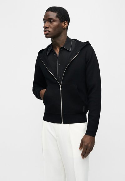 Άνδρας που φοράει μαύρη ζακέτα με φερμουάρ πάνω από μαύρο πουκάμισο με γιακά και άσπρο παντελόνι, στέκεται με το ένα χέρι στην τσέπη, κοιτάζοντας προς τα αριστερά.