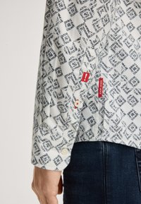 Witte shirt met blauw geometrisch patroon, knoopmanchet en twee rode labels. De stof lijkt lichtgewicht met een gladde textuur.