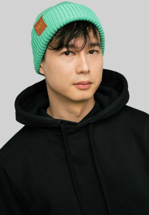 Jasnozielona, prążkowana czapka beanie z brązową skórzaną naszywką, noszona z czarną bluzą z kapturem. Prosty design, dopasowana, o teksturowanej powierzchni.