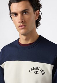 Giovane uomo con capelli ricci e scuri che indossa una felpa Champion blu navy e bianca con logo ricamato sul petto, che guarda dritto davanti a sé.