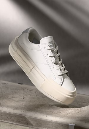 Sneaker bianca a bassa altezza con suola spessa beige, lacci frontali e cuciture sottili, esposta su una superficie in cemento con sfondo di ombra morbida.