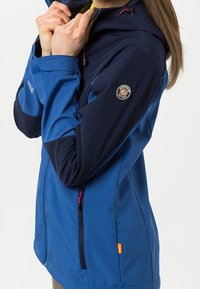Femme ajustant le col d'une veste softshell bleu et marine avec écussons de marque et poches zippées.