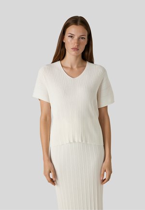 Jonge vrouw met steil bruin haar die een wit geribd kortemouwen top en bijpassende rok draagt, staand tegen een effen grijze achtergrond.