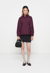 Bordeaux blouse met opstaande mouwen, gecombineerd met een marineblauwe geruite rok. Accessoires zijn een zwarte handtas en lichtbeige sokken.