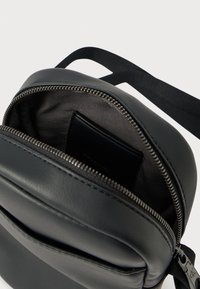 Černá kožená taška s hlavní přihrádkou na zip, která má hladkou texturu a jemnou vnitřní kapsu. Součástí je nastavitelný popruh.