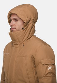 Brun isoleret jakke med høj krave og justerbar hætte. Har logo-patch på ærmet og snoretræk til tilpasning af pasform.