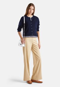 Kobieta w granatowym kardiganie noszącym paski nad białą koszulką, w beżowych szerokich spodniach i brązowych płaskich butach, trzymająca małą białą torbę na ramieniu z łańcuszkiem.