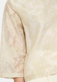 Blusa beige con delicati ricami floreali, texture semi-trasparente, maniche a tre quarti e una finitura di tessuto liscia e leggera.