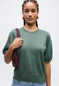 Žena s vrkôčikmi, ktorá má na sebe zlaté kruhové náušnice, zelený sveter s krátkym rukávom, drží vínovočervenú pletenú koženú kabelku na rameno.