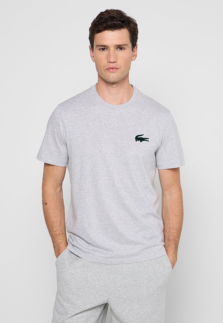 Homme debout avec les mains dans les poches portant un t-shirt gris clair et un pantalon assorti, avec un petit logo de crocodile noir sur la poitrine.