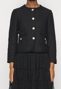 Černá texturovaná bunda s kulatým výstřihem, zlatým zapínáním na knoflíky a dvěma předními kapsami. Má jemně třpytivou tkaninu.