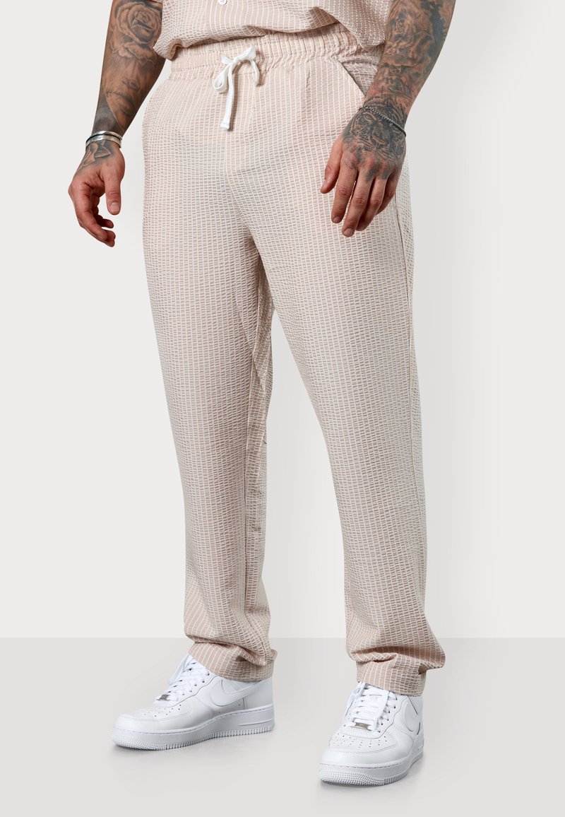 Light beige patterned joggers with an elastic waistband and drawstring, paired with white sneakers. Features a textured grid design.