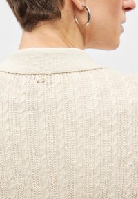 Μπεζ πλεκτή ζακέτα με ριμπ υφή, γιακά και μικρό μεταλλικό διακοσμητικό κουμπί στο πίσω μέρος.