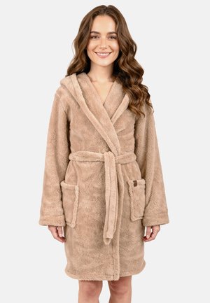 Jonge vrouw met lang golvend haar die lacht, gekleed in een beige zachte badjas met capuchon, zakken en een gestrikte riem, staand tegen een effen achtergrond.